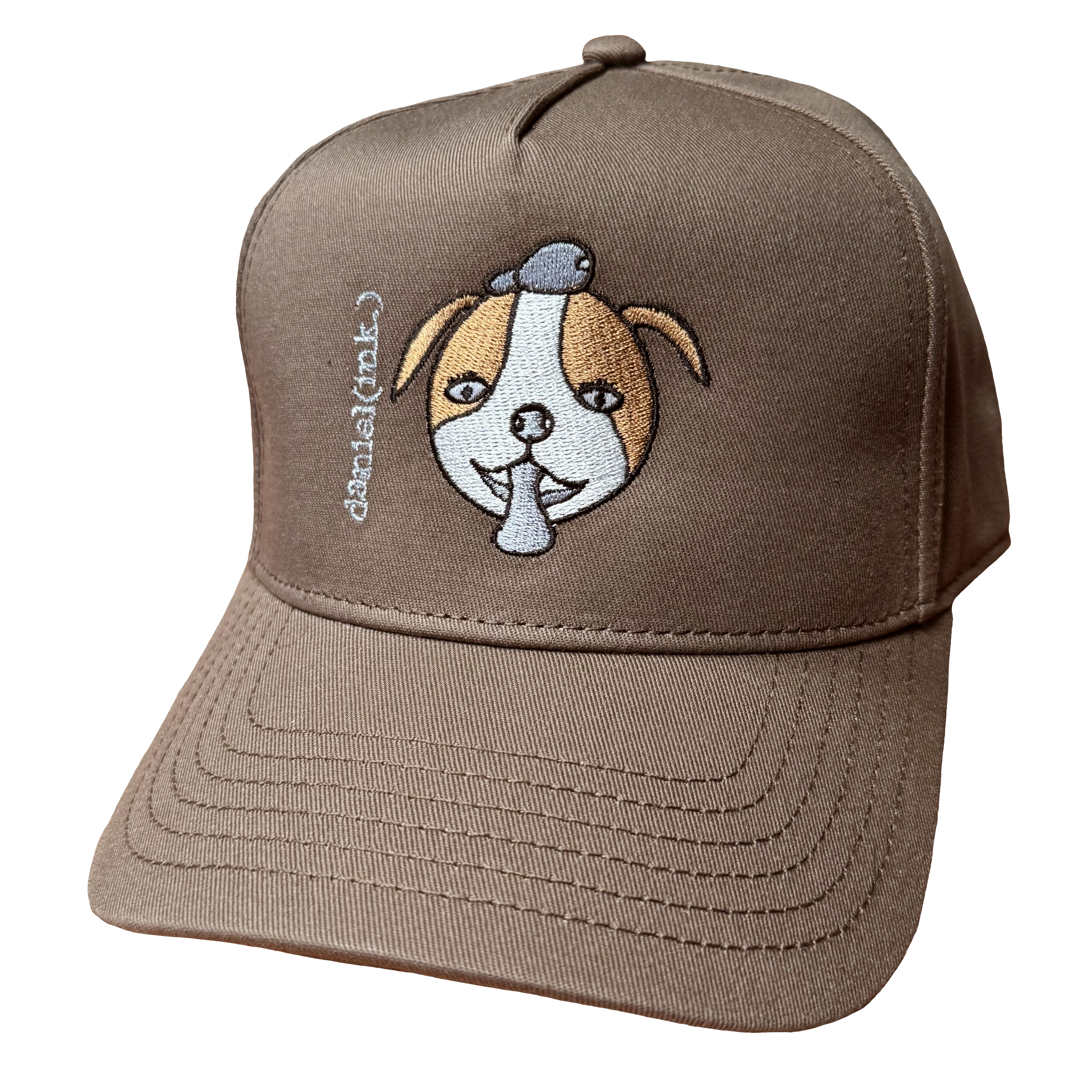Walnut snapback cap featuring an embroidered image of a dog's face in brown and white. The dog has a round head, floppy ears, wide eyes, and its tongue is sticking out. The name daniel(ink.) is embroidered in silver thread next to the dog's face.