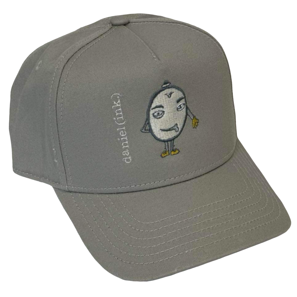 Storm snapback cap featuring an embroidered image of a humanoid egg man in silver. The egg man is wearing a silver bowler hat, gold gloves, and gold shoes, and is sticking his tongue out. The name daniel(ink.) is embroidered in silver thread next to the egg man's face.