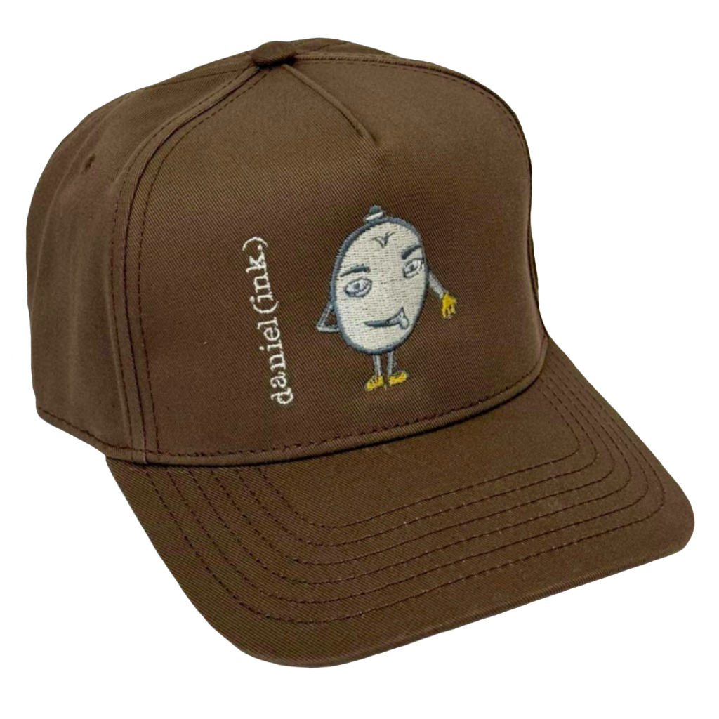 Walnut snapback cap featuring an embroidered image of a humanoid egg man in silver. The egg man is wearing a silver bowler hat, gold gloves, and gold shoes, and is sticking his tongue out. The name daniel(ink.) is embroidered in silver thread next to the egg man's face.